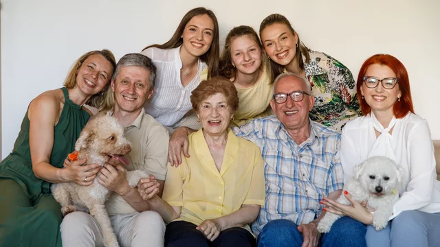 Eine fröhliche Familiengruppe posiert zusammen auf einer Couch. Es sind fünf Frauen und drei Männer, darunter ältere und jüngere Personen. Einige halten kleine Hunde in den Armen. Alle lächeln und strahlen Freude aus. Die Umgebung ist hell und einfach gehalten.