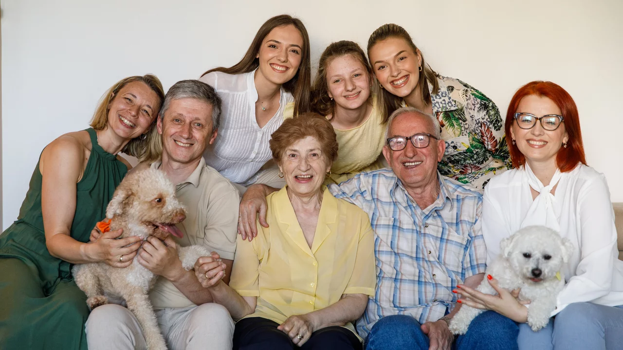 Eine fröhliche Familiengruppe posiert zusammen auf einer Couch. Es sind fünf Frauen und drei Männer, darunter ältere und jüngere Personen. Einige halten kleine Hunde in den Armen. Alle lächeln und strahlen Freude aus. Die Umgebung ist hell und einfach gehalten.