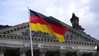 Eine deutsche Flagge weht vor dem Reichstagsgebäude in Berlin. Der Schriftzug „Dem Deutschen Volke“ ist über dem Eingang sichtbar. Der Himmel ist bewölkt, was eine dramatische Kulisse schafft.