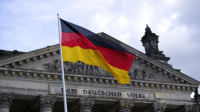 Eine deutsche Flagge weht vor dem Reichstagsgebäude in Berlin. Der Schriftzug „Dem Deutschen Volke“ ist über dem Eingang sichtbar. Der Himmel ist bewölkt, was eine dramatische Kulisse schafft.