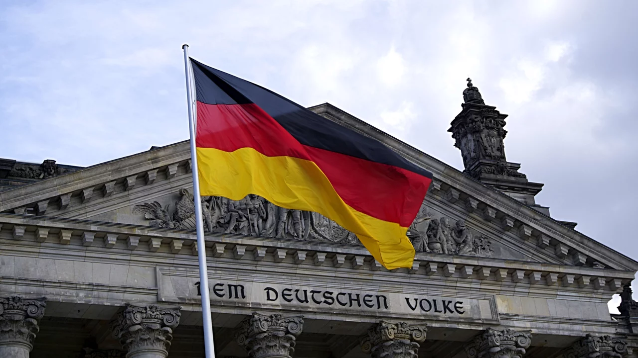 Eine deutsche Flagge weht vor dem Reichstagsgebäude in Berlin. Der Schriftzug „Dem Deutschen Volke“ ist über dem Eingang sichtbar. Der Himmel ist bewölkt, was eine dramatische Kulisse schafft.