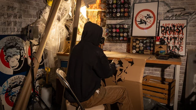 Ein Künstler in einem dunklen Raum arbeitet an einer Kartonfigur. Er trägt einen Kapuzenpullover und sitzt an einem Tisch mit Spraydosen und Kunstwerken im Hintergrund. An der Wand sind verschiedene Graffiti und Zeichnungen sichtbar, die urbanen Kunststil widerspiegeln.