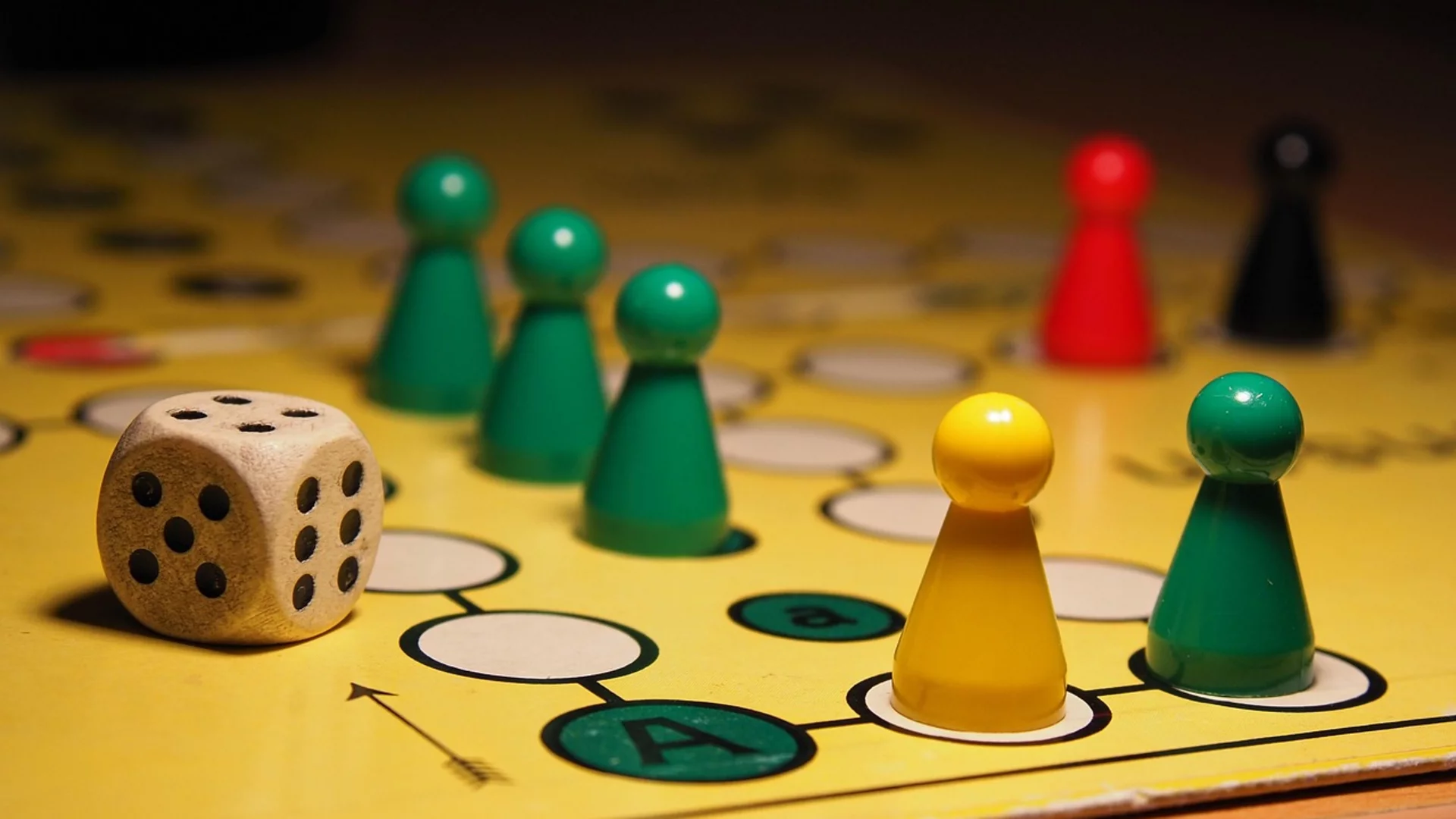 Auf einem Spielbrett sind mehrere farbige Spielfiguren (grün, rot, gelb, schwarz) positioniert. Ein Holzspielwürfel liegt daneben. Die Szenerie vermittelt die Atmosphäre eines Brettspiels, das gerade gespielt wird.