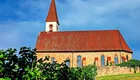 Eine Kirche mit einem spitzen roten Dach und einem Glockenturm steht vor einem blauen Himmel. Das Gebäude ist aus Stein und hat mehrere Fenster. Im Vordergrund wachsen grüne Weinreben.