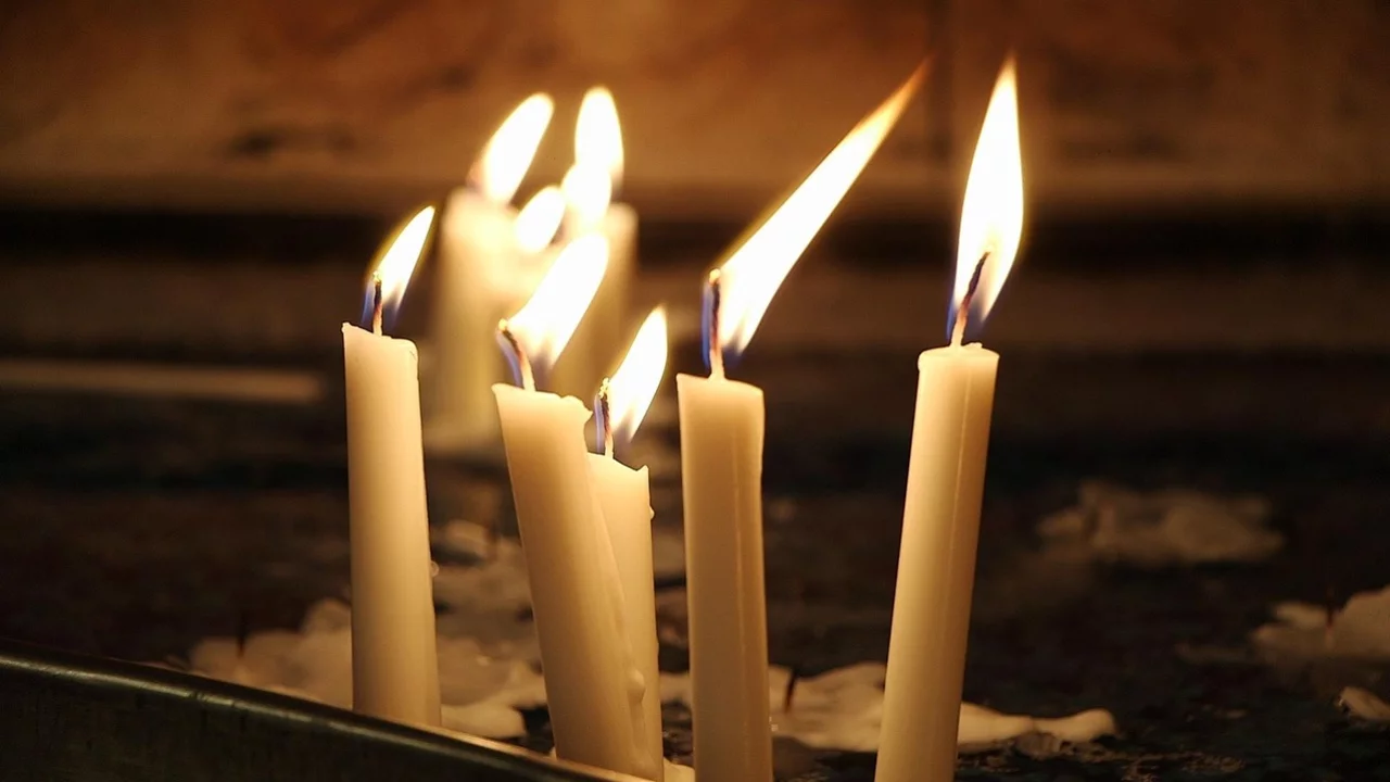 Sechs brennende Kerzen stehen auf einer Unterlage, ihre Flammen flackern sanft im Dunkeln. Die Kerzen sind beige und erzeugen eine ruhige, besinnliche Atmosphäre.