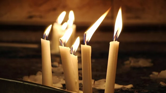 Sechs brennende Kerzen stehen auf einer Unterlage, ihre Flammen flackern sanft im Dunkeln. Die Kerzen sind beige und erzeugen eine ruhige, besinnliche Atmosphäre.