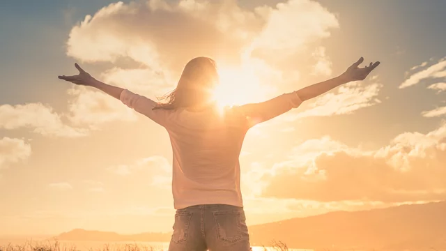 Eine Person steht mit ausgebreiteten Armen vor einem Sonnenuntergang. Das Licht der Sonne strahlt hinter ihr hervor, während weiche Wolken den Himmel schmücken. Die Szene vermittelt ein Gefühl von Freiheit und Freude in der Natur.