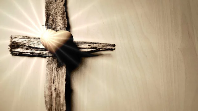 Ein hölzernes Kreuz mit einem goldenen Herz in der Mitte. Lichtstrahlen scheinen von hinten auf das Kreuz, was eine warme und symbolische Atmosphäre schafft.