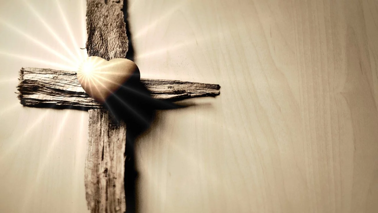 Ein hölzernes Kreuz mit einem goldenen Herz in der Mitte. Lichtstrahlen scheinen von hinten auf das Kreuz, was eine warme und symbolische Atmosphäre schafft.