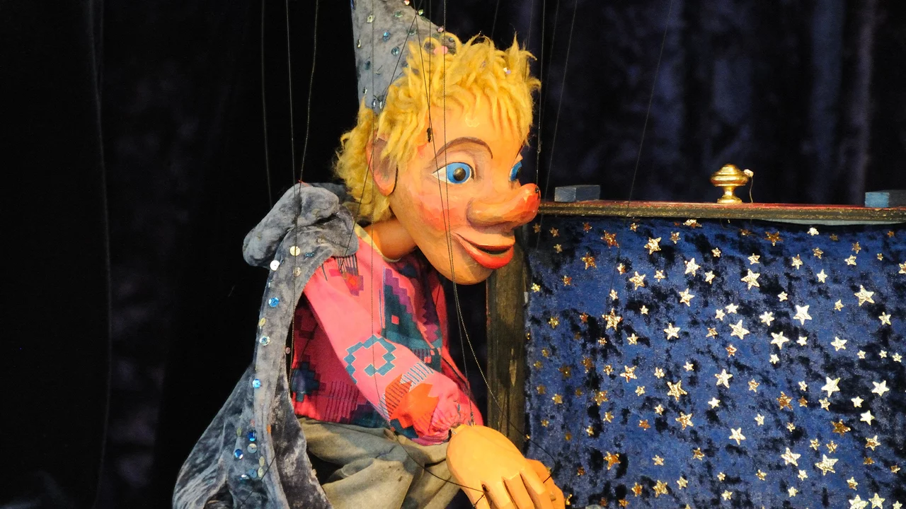 Eine bunte Marionette mit blonden Haaren und roten Lippen sitzt vor einer glänzenden Truhe, die mit Sternen verziert ist. Sie trägt einen glitzernden Umhang und einen spitzen Hut und scheint gebannt auf etwas zu schauen.