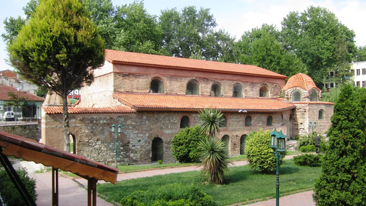 Eine historische, steinerne Struktur mit einem roten Ziegeldach, umgeben von hohen Bäumen und Grünflächen. Teile des Gebäudes sind sichtbar mit Fenstern und Bögen. Es befindet sich in einem parkähnlichen Gelände mit anderen Pflanzen.