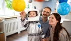 Ein kleines Kind in einem silbernen Astronautenanzug greift nach schwebenden Planeten in einem Raum. Die Eltern betrachten es lächelnd. Die Atmosphäre ist freundlich und spielerisch, ein Moment des gemeinsamen Entdeckens der Welt des Weltraums.