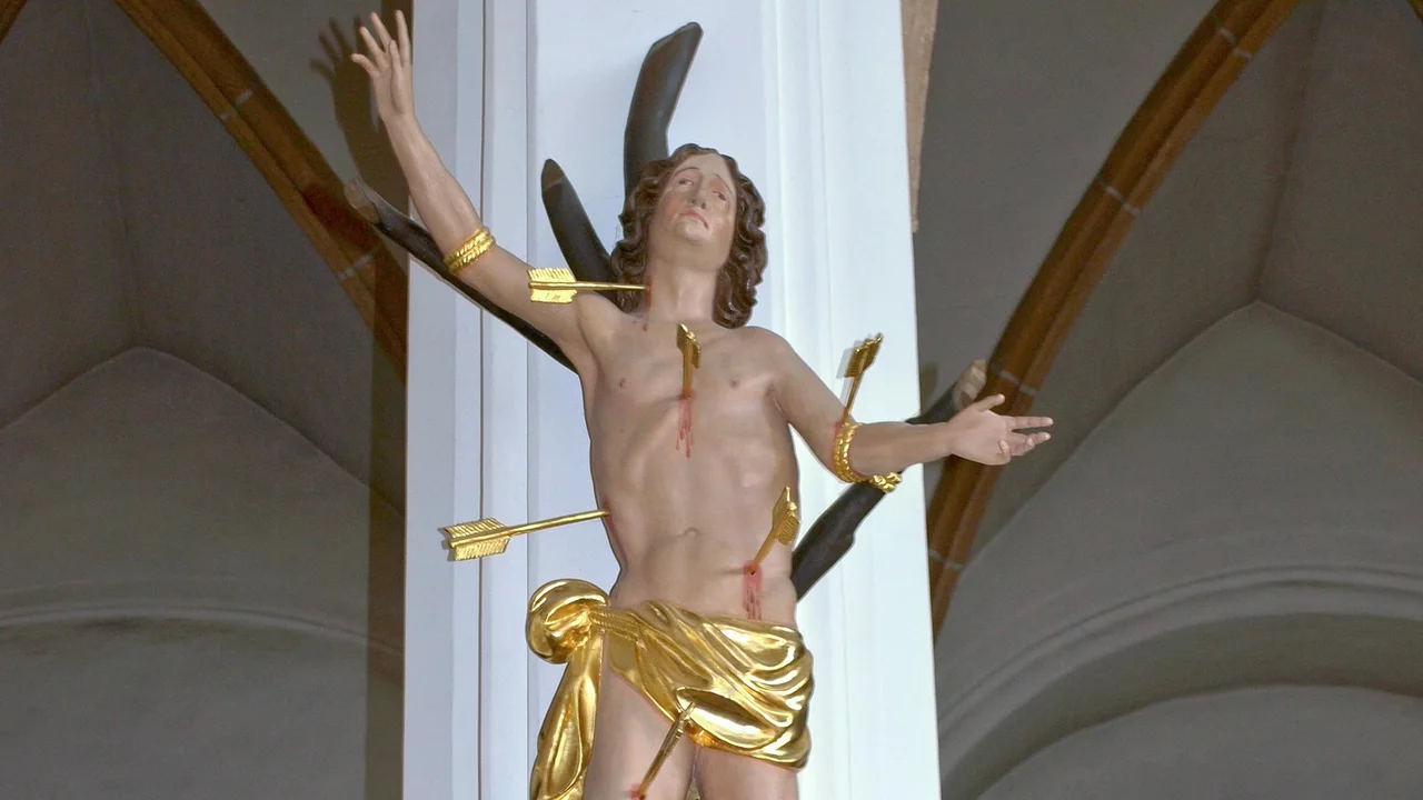 Eine Statue von Jesus, der am Kreuz hängt, mit ausgebreiteten Armen. Er trägt nur ein Lendentuch und hat mehrere Pfeile im Körper. Die Figur ist kunstvoll in gold und braun gestaltet und befindet sich an einer Säule in einer Kirche.