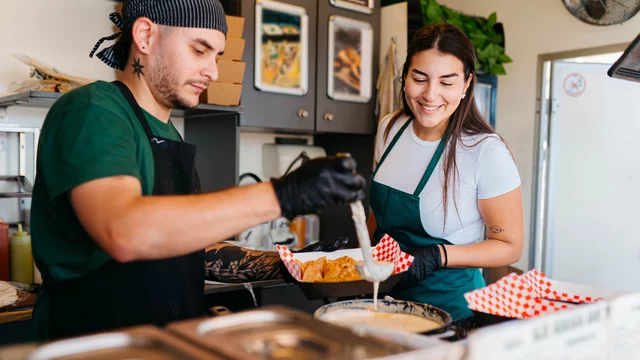 Ein Koch und eine Mitarbeiterin in einem Restaurant bereiten Essen zu. Der Koch trägt Handschuhe und schöpft mit einer Kelle, während die Mitarbeiterin lächelnd zusieht. Im Hintergrund sind verschiedene Lebensmittel und Küchenutensilien sichtbar.