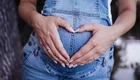 Eine schwangere Person hält ihren Bauch mit beiden Händen, die eine Herzform bilden. Sie trägt einen blauen Overall und steht in einer natürlichen Umgebung. Der Fokus liegt auf der Darstellung der Schwangerschaft und der emotionalen Verbindung.