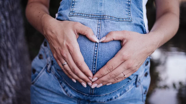 Eine schwangere Person hält ihren Bauch mit beiden Händen, die eine Herzform bilden. Sie trägt einen blauen Overall und steht in einer natürlichen Umgebung. Der Fokus liegt auf der Darstellung der Schwangerschaft und der emotionalen Verbindung.