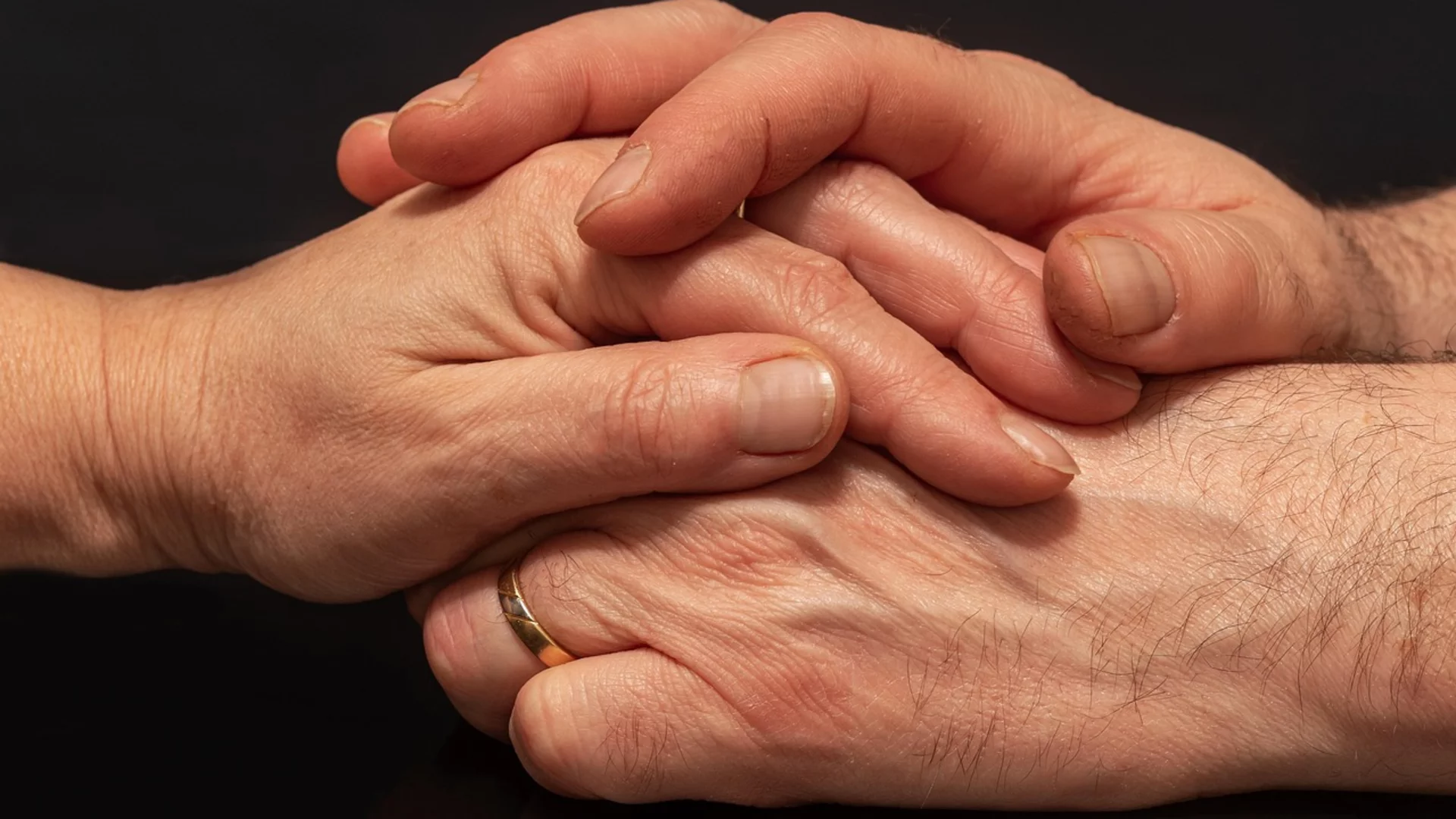 Zwei Hände liegen übereinander, eine mit einem Ring. Die Hände sind sanft und symbolisieren Nähe und Verbundenheit. Der Hintergrund ist dunkel, was die Hände hervorhebt.