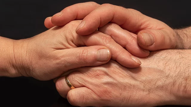 Zwei Hände liegen übereinander, eine mit einem Ring. Die Hände sind sanft und symbolisieren Nähe und Verbundenheit. Der Hintergrund ist dunkel, was die Hände hervorhebt.