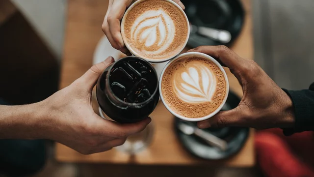 Drei Hände halten Tassen mit Getränken, darunter zwei Cappuccinos mit Herz-Muster auf der Oberfläche und ein Glas mit einem dunklen Getränk. Die Szene vermittelt ein Gefühl von Gemeinsamkeit und Genuss in einer gemütlichen Umgebung.
