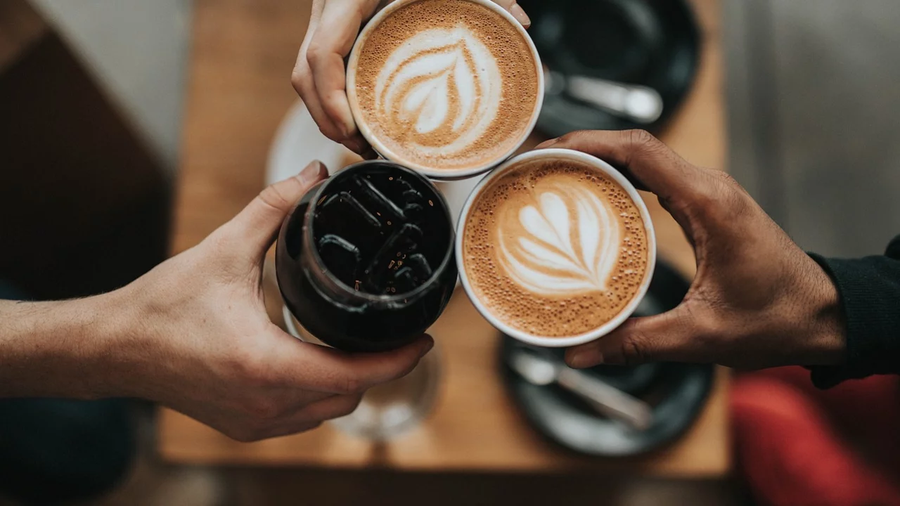 Drei Hände halten Tassen mit Getränken, darunter zwei Cappuccinos mit Herz-Muster auf der Oberfläche und ein Glas mit einem dunklen Getränk. Die Szene vermittelt ein Gefühl von Gemeinsamkeit und Genuss in einer gemütlichen Umgebung.