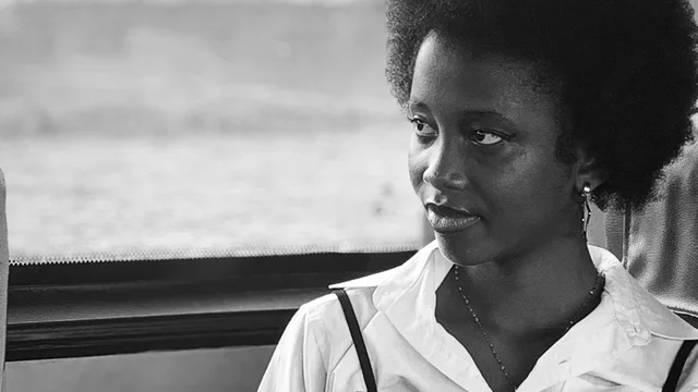 Eine junge schwarze Frau mit lockigem Haar sitzt in einem Bus. Sie trägt ein weißes Hemd und hat einen nachdenklichen Ausdruck. Der Hintergrund ist unscharf, mit einem Fenster, das nur leicht sichtbar ist.