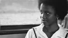 Eine junge schwarze Frau mit lockigem Haar sitzt in einem Bus. Sie trägt ein weißes Hemd und hat einen nachdenklichen Ausdruck. Der Hintergrund ist unscharf, mit einem Fenster, das nur leicht sichtbar ist.