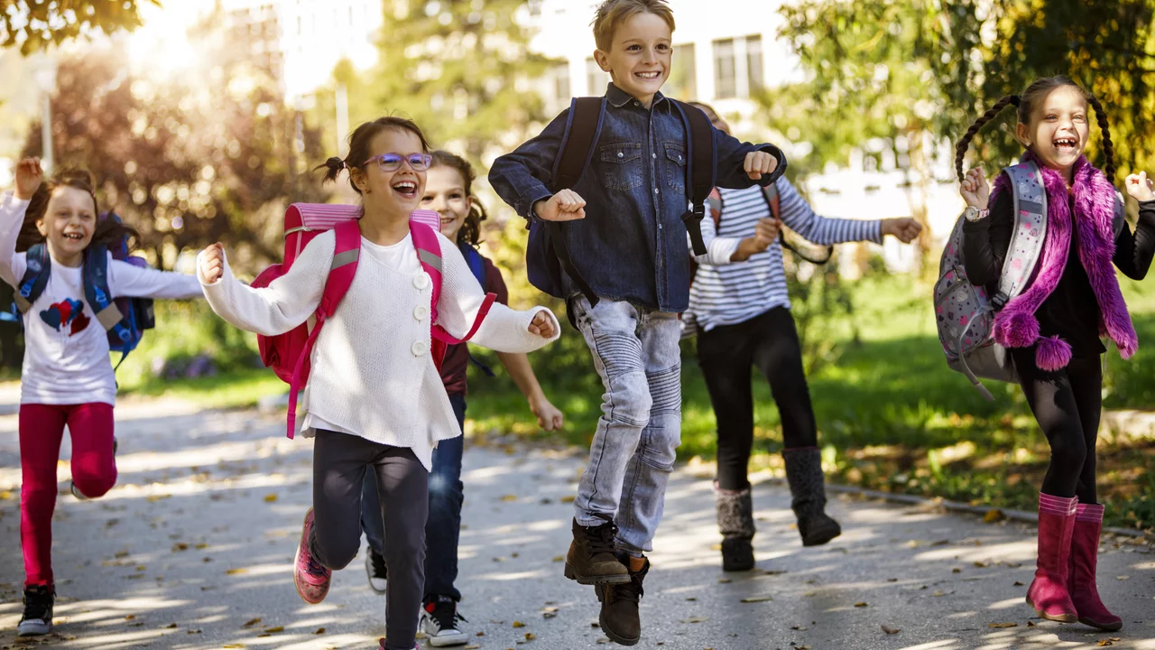 Eine Gruppe von fröhlichen Kindern läuft lachend auf einem Weg im Freien. Sie tragen Schulranzen und sind in lässiger Kleidung. Die Sonne scheint, und das Bild vermittelt eine Stimmung von Freude und Unbeschwertheit.
