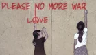 Zwei Mädchen stehen vor einer hellen Wand . Die größere von beiden steht am rechten Rand und schreibt mit roter Farbe "Pleace no more war" an die Wand. Das kleinere Mädchen schaut zu ihr hin und zeigt mit ihrem linken ausgestreckten Arm auf das Wort "Love" über ihr.