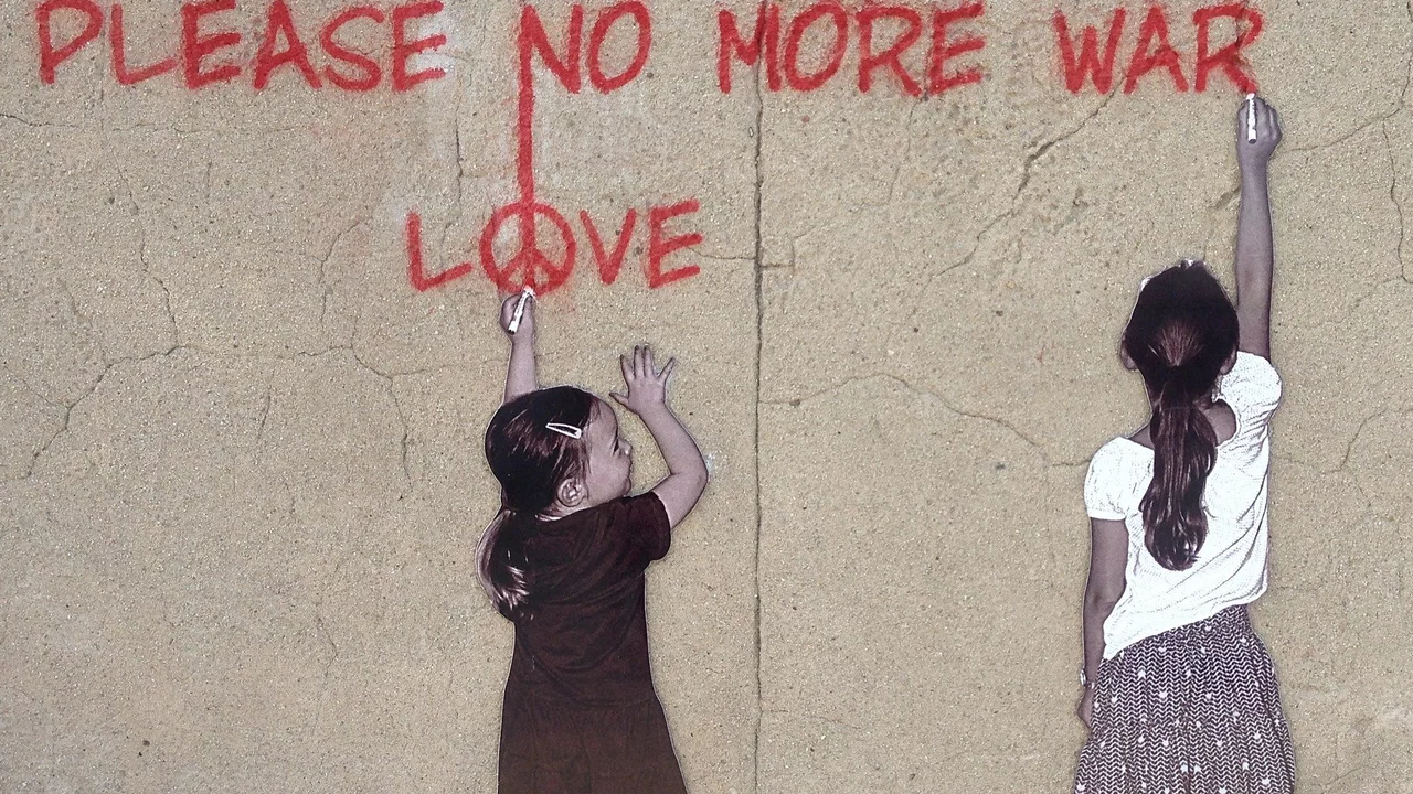 Zwei Mädchen stehen vor einer hellen Wand . Die größere von beiden steht am rechten Rand und schreibt mit roter Farbe "Pleace no more war" an die Wand. Das kleinere Mädchen schaut zu ihr hin und zeigt mit ihrem linken ausgestreckten Arm auf das Wort "Love" über ihr.