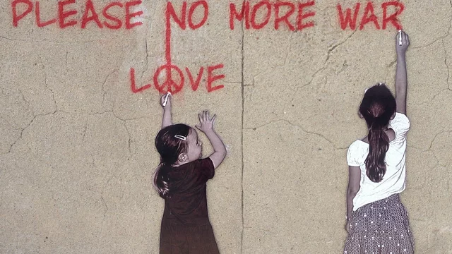 Zwei Mädchen stehen vor einer hellen Wand . Die größere von beiden steht am rechten Rand und schreibt mit roter Farbe "Pleace no more war" an die Wand. Das kleinere Mädchen schaut zu ihr hin und zeigt mit ihrem linken ausgestreckten Arm auf das Wort "Love" über ihr.