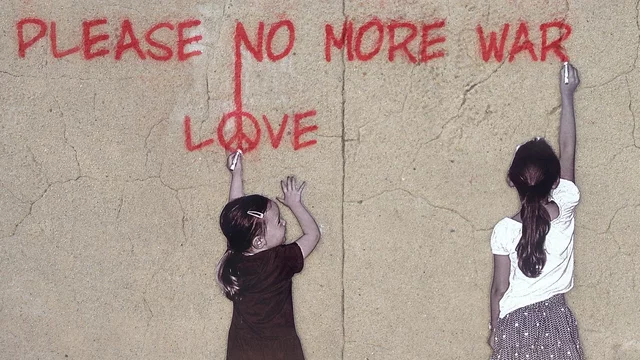Zwei Mädchen stehen vor einer hellen Wand . Die größere von beiden steht am rechten Rand und schreibt mit roter Farbe "Pleace no more war" an die Wand. Das kleinere Mädchen schaut zu ihr hin und zeigt mit ihrem linken ausgestreckten Arm auf das Wort "Love" über ihr.
