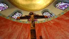 Ein Holz-Kreuz mit der Darstellung von Jesus Christus ist im Mittelpunkt des Bildes zu sehen. Der Hintergrund zeigt bunte Glasfenster und eine goldene Decke. Die Wand ist in rötlichen Farbtönen gehalten, was eine ruhige und meditative Atmosphäre schafft.