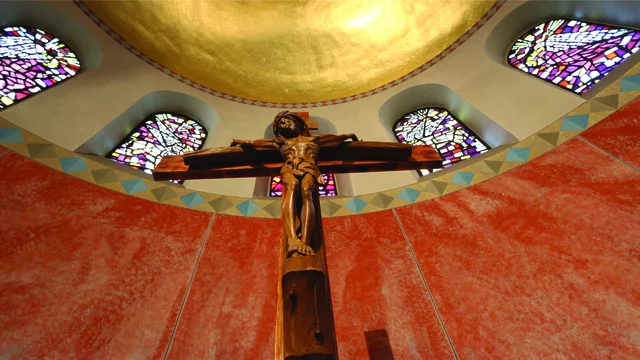 Ein Holz-Kreuz mit der Darstellung von Jesus Christus ist im Mittelpunkt des Bildes zu sehen. Der Hintergrund zeigt bunte Glasfenster und eine goldene Decke. Die Wand ist in rötlichen Farbtönen gehalten, was eine ruhige und meditative Atmosphäre schafft.