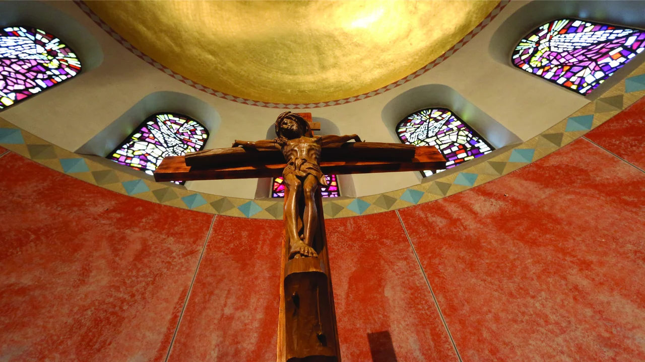Ein Holz-Kreuz mit der Darstellung von Jesus Christus ist im Mittelpunkt des Bildes zu sehen. Der Hintergrund zeigt bunte Glasfenster und eine goldene Decke. Die Wand ist in rötlichen Farbtönen gehalten, was eine ruhige und meditative Atmosphäre schafft.