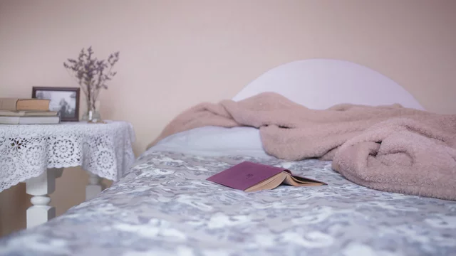 Ein Bett mit einer einladenden, gestreiften Bettdecke ist im Vordergrund zu sehen. Auf dem Bett liegt ein aufgeschlagenes Buch. Neben dem Bett steht ein Tisch mit einer Decke und einigen Büchern sowie einer kleinen Vase mit getrockneten Blumen.
