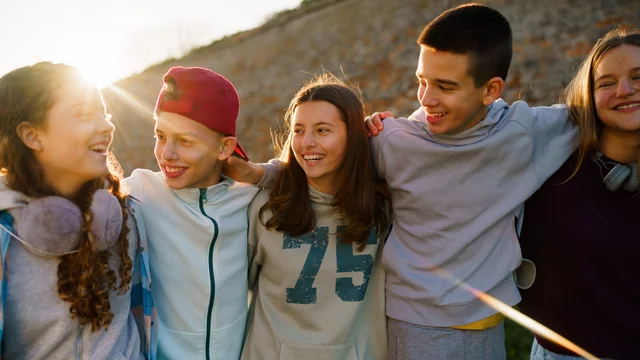 Eine Gruppe von fünf Jugendlichen steht in einer Reihe und lächelt miteinander. Sie wirken fröhlich und entspannt, umarmt oder legt die Arme über die Schultern des jeweils anderen. Im Hintergrund ist eine natürliche Landschaft bei Sonnenuntergang zu sehen.