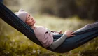 Eine entspannte Frau liegt in einer Hängematte, mit geschlossenen Augen und einem sanften Lächeln. Sie trägt einen beigen Hut und eine weiche Jacke. Die Umgebung scheint ruhig und grün, ideal zum Entspannen und Ausruhen.