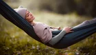 Eine entspannte Frau liegt in einer Hängematte, mit geschlossenen Augen und einem sanften Lächeln. Sie trägt einen beigen Hut und eine weiche Jacke. Die Umgebung scheint ruhig und grün, ideal zum Entspannen und Ausruhen.