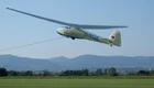 Ein Segelflugzeug mit der Kennung D-7138 ist in der Luft, während es durch eine Leine mit einem Bodentransportmittel verbunden ist. Der Hintergrund zeigt eine hügelige Landschaft unter klarem Himmel.