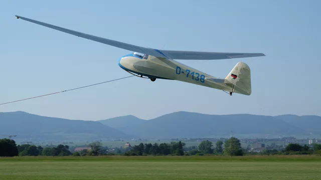 Ein Segelflugzeug mit der Kennung D-7138 ist in der Luft, während es durch eine Leine mit einem Bodentransportmittel verbunden ist. Der Hintergrund zeigt eine hügelige Landschaft unter klarem Himmel.