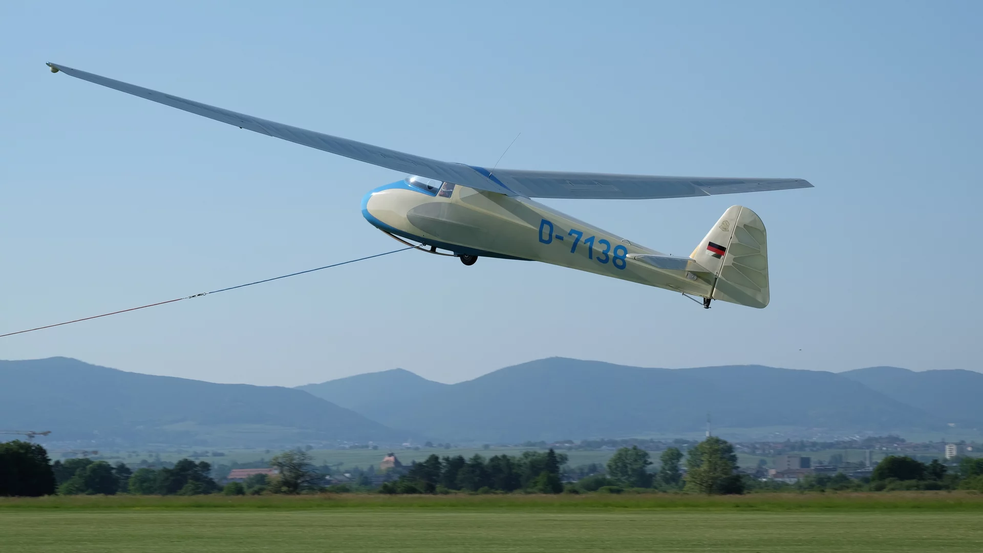 Ein Segelflugzeug mit der Kennung D-7138 ist in der Luft, während es durch eine Leine mit einem Bodentransportmittel verbunden ist. Der Hintergrund zeigt eine hügelige Landschaft unter klarem Himmel.