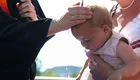 Ein Kleinkind in einem zartrosa Kleidchen wird von einer Frau über eine Taufschale gehalten. Davor steht eine Pfarrperson in schwarzem Talar und schöpft mit der Hand etwas Wasser auf den Kopf des Kindes. Die Szene findet im Freien statt.