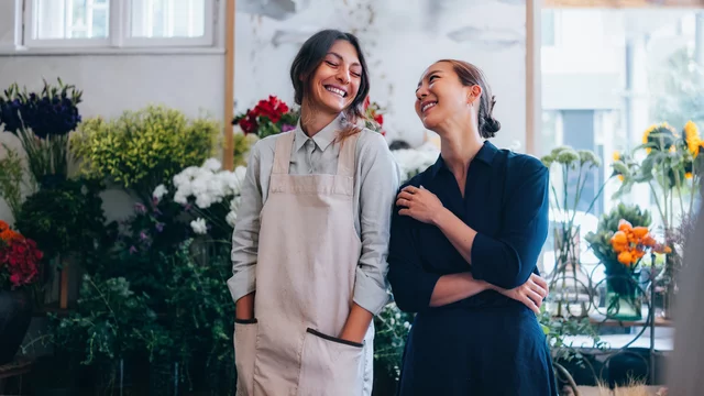 Zwei Frauen stehen in einem Blumenladen, umgeben von bunten Blumenarrangements. Sie lächeln sich an und scheinen eine freundliche Unterhaltung zu führen. Eine der Frauen trägt eine Schürze, die andere ein dunkelblaues Kleid. 