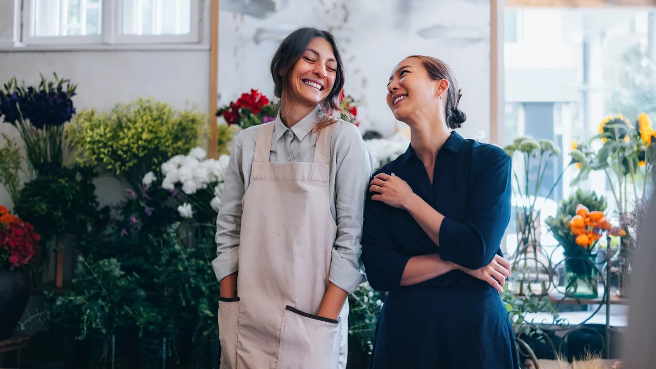 Zwei Frauen stehen in einem Blumenladen, umgeben von bunten Blumenarrangements. Sie lächeln sich an und scheinen eine freundliche Unterhaltung zu führen. Eine der Frauen trägt eine Schürze, die andere ein dunkelblaues Kleid. 