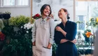 Zwei Frauen stehen in einem Blumenladen, umgeben von bunten Blumenarrangements. Sie lächeln sich an und scheinen eine freundliche Unterhaltung zu führen. Eine der Frauen trägt eine Schürze, die andere ein dunkelblaues Kleid. 