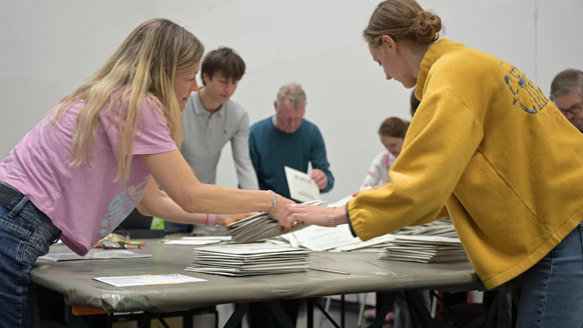 Eine Gruppe von Menschen arbeitet zusammen an einem Tisch, um Papiere oder Dokumente zu sortieren und zu stapeln. Zwei Personen im Vordergrund greifen nach den gleichen Blättern, während weitere Personen im Hintergrund beschäftigt sind. Die Atmosphäre ist kooperativ und engagiert.
