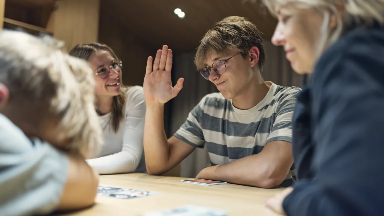 Eine Gruppe von vier Personen sitzt um einen Tisch. Ein junger Mann hebt die Hand, während eine Frau lächelt. Ein anderer junger Mann liegt mit dem Kopf auf dem Tisch, und eine weitere Frau schaut aufmerksam. Es scheint, dass sie zusammen ein Spiel spielen.