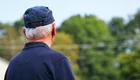 Ein älterer Mann mit weißen Haaren trägt eine blaue Mütze und ein dunkles Hemd. Er steht mit dem Rücken zur Kamera und schaut in eine grüne, bewaldete Landschaft. Der Himmel ist blau mit wenigen Wolken.