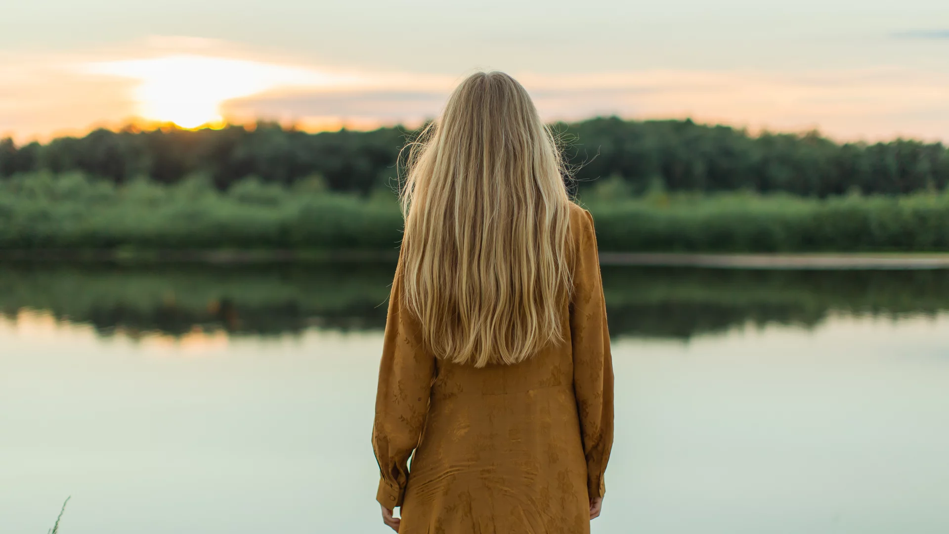 Eine Person mit langen Haaren steht am Ufer eines ruhigen Wassers und blickt auf einen Sonnenuntergang. Der Himmel ist in sanften Farben erleuchtet, während im Hintergrund grüne Pflanzen zu sehen sind. Die Szene vermittelt ein Gefühl von Ruhe und Naturverbundenheit.