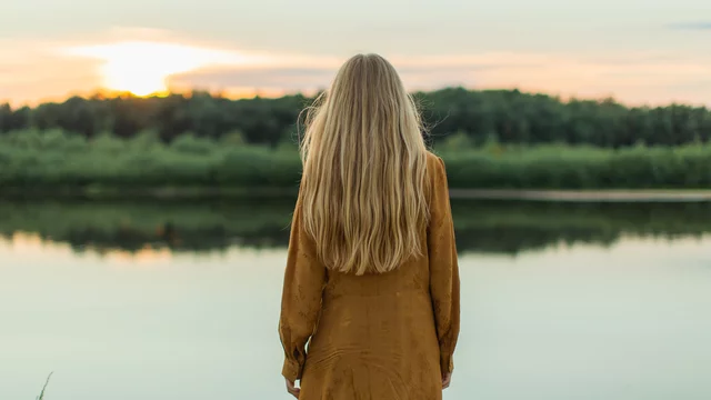 Eine Person mit langen Haaren steht am Ufer eines ruhigen Wassers und blickt auf einen Sonnenuntergang. Der Himmel ist in sanften Farben erleuchtet, während im Hintergrund grüne Pflanzen zu sehen sind. Die Szene vermittelt ein Gefühl von Ruhe und Naturverbundenheit.